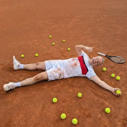 Achim Seemann liegt lachend und erschöpft auf dem Tennisplatz, umgeben von Tennisbällen