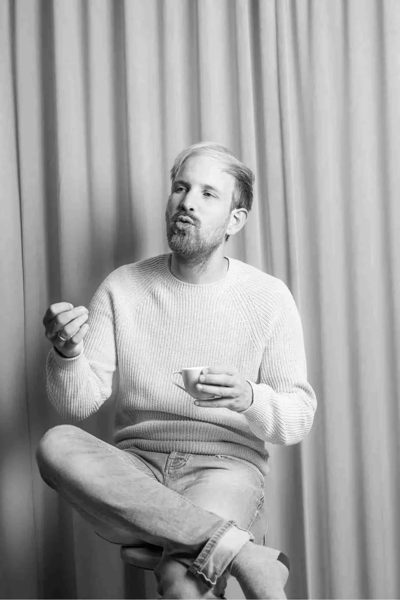 Foto von Rutger Bregman, der auf einem Hocker mit überschlagenen Beinen, mit einer Tasse in der Hand gestikuliert. 