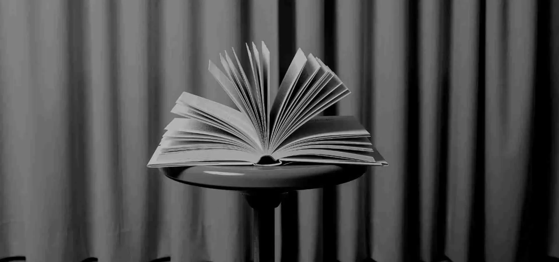 Foto von einem aufgeschlagenen Buch mit aufgeblätterten Seiten auf einem Hocker vor einem Vorhang. 