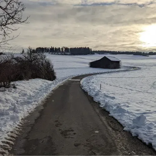 Verschneite Landstraße führt zu einer Scheune bei Sonnenuntergang